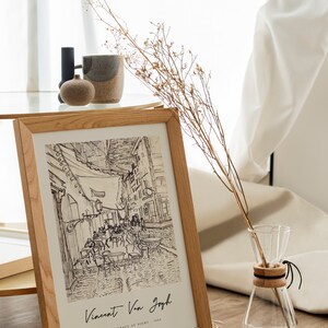 May include: A framed black and white drawing of a cafe terrace at night by Vincent Van Gogh. The drawing is in a light wood frame and is sitting on a table with a vase of dried flowers.