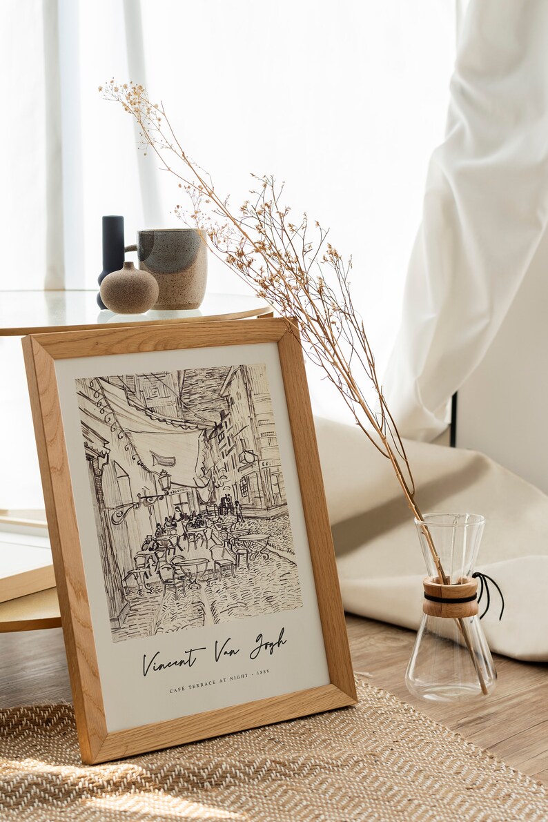 May include: A framed black and white drawing of a cafe terrace at night by Vincent Van Gogh. The drawing is in a light wood frame and is sitting on a table with a vase of dried flowers.