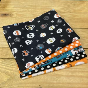 May include: A stack of five folded fabric pieces featuring various patterns. The top piece is black with white and orange skull motifs. Below are orange with white polka dots, blue with skeleton graphics, black with white polka dots, and orange with ghosts.