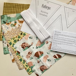 May include: A collection of fabric pieces with various patterns, including pigs, chickens, and a gingham design. A white ribbon and a bunting template with instructions are also present. The fabrics are in shades of green, yellow, and brown.
