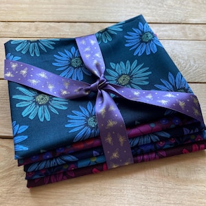 May include: A bundle of fabric squares in shades of blue and purple, decorated with floral patterns. The fabric is tied with a purple ribbon with gold bee detailing. The stack includes various floral designs, creating a visually appealing textile bundle.