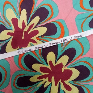 May include: Fabric with a large floral pattern in shades of pink, teal, yellow, orange, and burgundy. The fabric has a repeating design of stylized flowers. A white ribbon across the fabric reads "Brandon Mably for Rowan. #BM 11 Flora."