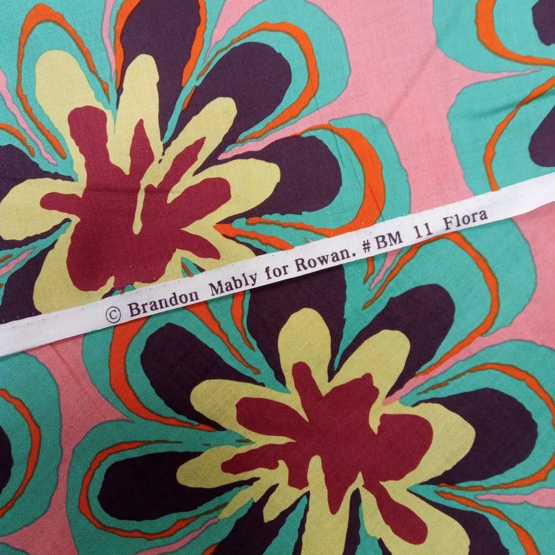 May include: Fabric with a large floral pattern in shades of pink, teal, yellow, orange, and burgundy. The fabric has a repeating design of stylized flowers. A white ribbon across the fabric reads "Brandon Mably for Rowan. #BM 11 Flora."