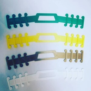 May include: Four different coloured plastic face mask extenders. The extenders are green, yellow, dark grey, and white. Each extender has a rectangular opening in the middle and five prongs on each end.
