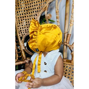 May include: A golden yellow satin baby bonnet with a ruffled brim and matching ties. The bonnet is worn by a baby wearing a white dress. The baby is also holding a matching satin scrunchie. The bonnet has a small logo on the side.