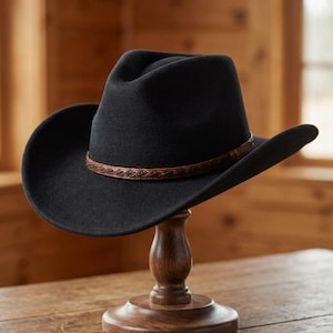 Può includere: Un cappello da cowboy in feltro nero con corona pizzicata e tesa larga e curva. Il cappello presenta una sottile fascia decorativa in pelle marrone attorno alla base della corona, ornata da un motivo scuro e complesso ripetuto al centro.