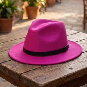 Puede incluir: Un sombrero fedora de color rosa intenso con una copa estructurada y un ala ancha y plana. El sombrero está acentuado con una banda negra lisa y sólida que rodea la base de la copa, creando un contraste marcado con el material de fieltro rosa.