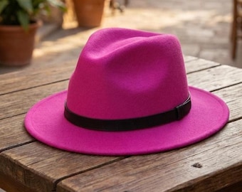 May include: A vibrant hot pink fedora hat featuring a structured crown and a wide, flat brim. The hat is accented with a smooth, solid black band that encircles the base of the crown, providing a distinct contrast to the bright pink felt material.
