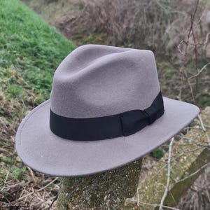 Puede incluir: Un sombrero fedora de color gris claro con una copa estructurada con pliegue central y ala ancha. El sombrero está adornado con una cinta de grosgrain negra lisa que rodea la base de la copa, terminada con un pequeño lazo lateral.