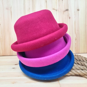 May include: Three felt hats stacked on top of each other. The top hat is a deep red, the middle hat is pink, and the bottom hat is blue.