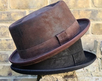 May include: Two fedora hats stacked on top of each other. The top hat is brown with a matching band and brim. The bottom hat is black with a black band and brim. The hats are resting on a stone surface in front of a brick wall.