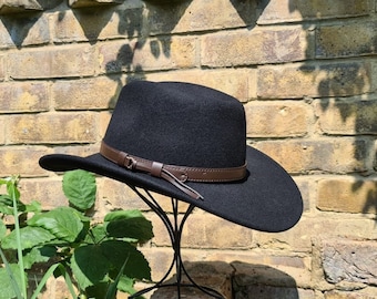 Op de afbeelding: Een zwarte cowboyhoed van vilt met een bruin leren band en een kwast. De hoed staat op een zwarte standaard.