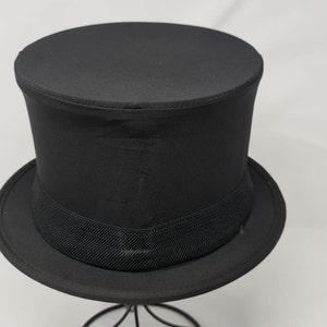 May include: A black top hat, featuring a wide brim and a black grosgrain ribbon band. The hat is displayed on a black metal stand against a white backdrop. The hat's crown is cylindrical.