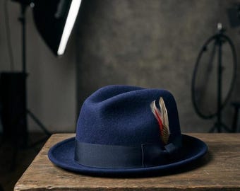 May include: A dark blue felt fedora hat featuring a matching dark blue grosgrain ribbon band and a decorative feather accent on the side. The hat has a pinched crown and a structured brim, showcasing a smooth, uniform texture throughout.