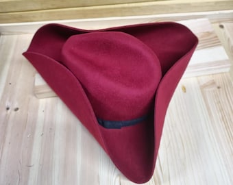 May include: A red felt tricorn hat with a black band around the crown. The hat is folded in half and lying on a wooden surface.