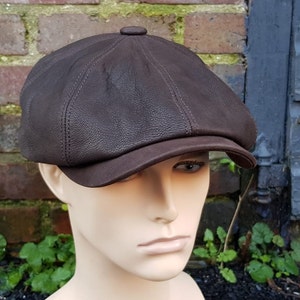 May include: A brown leather newsboy cap with a slightly curved brim. The cap has a textured surface and a button on the top.