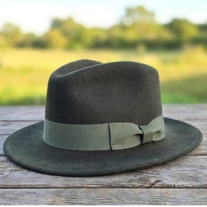 Op de afbeelding: Een donkergroene fedora hoed met een lichtgroene band en strik. De hoed is gemaakt van een viltachtig materiaal en staat op een verweerd houten oppervlak. De achtergrond is wazig en toont een veld en bomen.