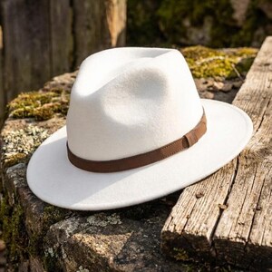 Könnte beinhalten: Ein weißer Fedora-Hut mit eingedrückter Krone und breiter Krempe. Der Hut verfügt über ein braunes Band, das die Basis der Krone umschließt und mit einer kleinen, passenden braunen Schlaufe befestigt ist. Die Oberfläche hat eine weiche Filzstruktur.