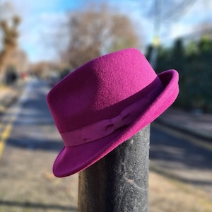 May include: A vibrant magenta felt fedora hat featuring a structured crown and a slightly upturned brim. The hat is accented with a matching magenta grosgrain ribbon band that includes a small, centred bow detail on the side of the crown.