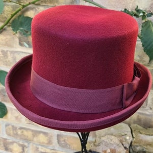 May include: A burgundy felt top hat with a matching burgundy ribbon band. The hat is on a black stand.