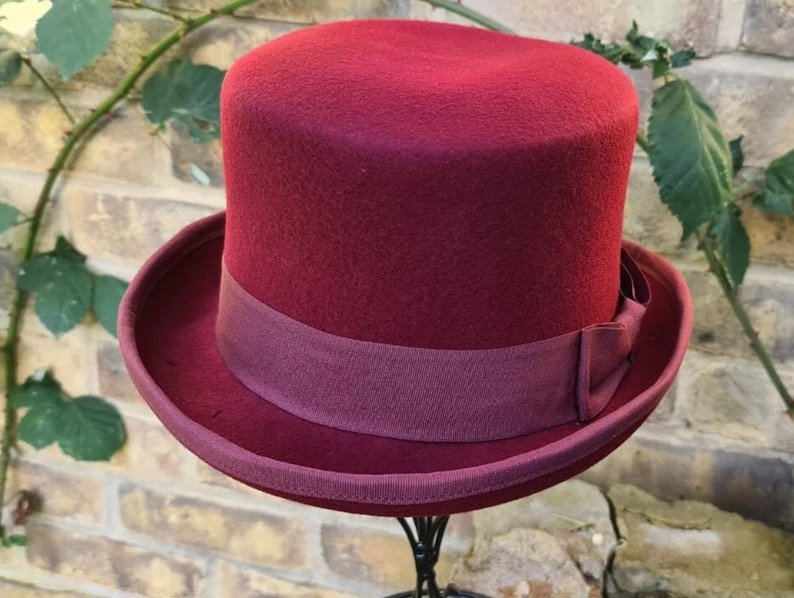 May include: A burgundy felt top hat with a matching burgundy ribbon band. The hat is on a black stand.