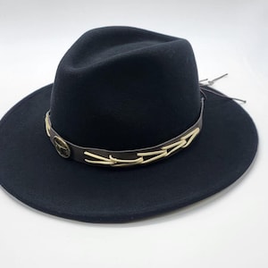 May include: A black fedora-style hat with a wide brim. The hat features a brown leather band with a decorative gold-colored emblem and cream-colored stitching. The hat is a classic accessory.