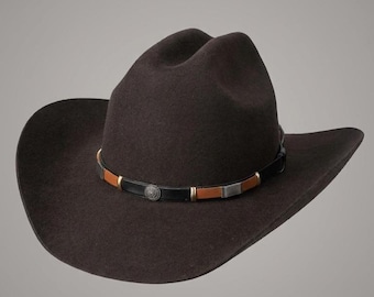 May include: A dark brown felt cowboy hat featuring a pinched crown and a wide, curved brim. The hat is accented with a decorative band composed of alternating black and tan leather segments, metallic hardware, and a circular silver-toned emblem.