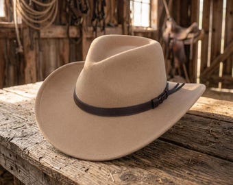 May include: A tan felt cowboy hat featuring a pinched crown and a wide, curved brim. The hat is accented with a thin, dark brown leather band that wraps around the base of the crown, secured with a small buckle and two short leather tails.