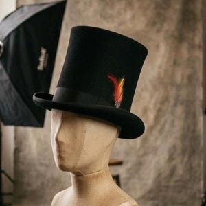 May include: A black top hat featuring a flat crown and a curved brim. The hat is adorned with a black grosgrain ribbon band and a single decorative feather accent in shades of red, orange, and yellow, positioned on the side of the crown.
