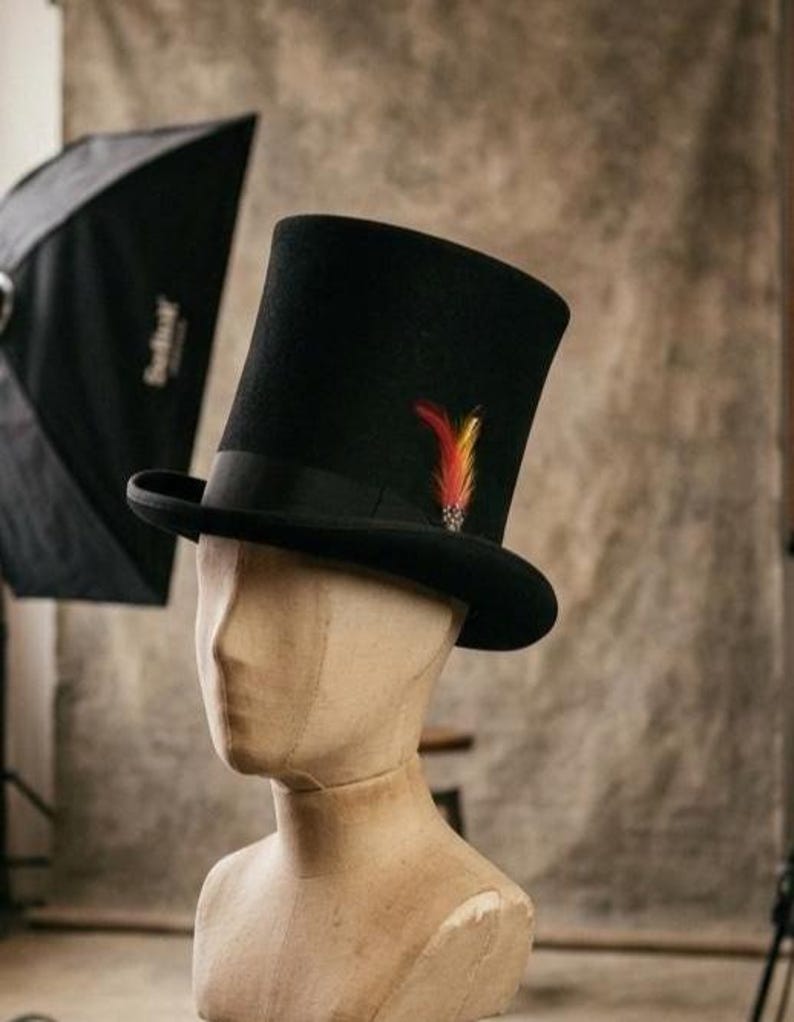 May include: A black top hat featuring a flat crown and a curved brim. The hat is adorned with a black grosgrain ribbon band and a single decorative feather accent in shades of red, orange, and yellow, positioned on the side of the crown.