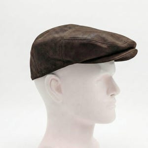 Könnte beinhalten: Eine dunkelbraune Leder-Flatcap mit abgerundeter Krone und kleinem, steifem Schirm. Die Kappe hat einen satten, erdigen Braunton und scheint aus weichem, strukturiertem Leder gefertigt zu sein. Präsentiert auf einem weißen Schaufensterkopf.