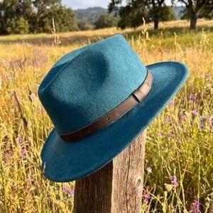 May include: A teal fedora hat with a structured crown and a wide brim. The hat features a dark brown leather band wrapped around the base of the crown, secured with a small metal buckle detail on the side. The material has a smooth, matte finish.