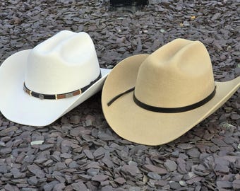 May include: Two cowboy hats, one white and one tan, featuring a creased crown and a wide brim. The white hat has a black band with metallic accents, while the tan hat features a simple black band. Both hats are constructed from a felt-like material.