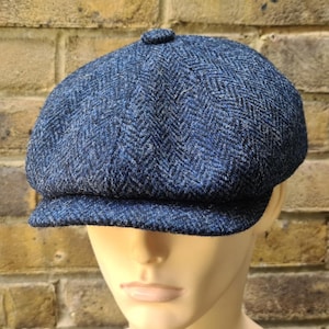 May include: A blue herringbone tweed newsboy cap with a rounded crown and a short, stiff brim. The cap is made of a textured fabric with a subtle pattern. The cap is displayed on a mannequin head.