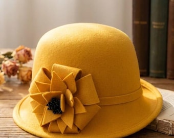 May include: A mustard yellow felt cloche hat featuring a rounded crown and a narrow brim. A large, layered fabric flower in the same mustard yellow hue is attached to the side, accented with a cluster of small, dark, bead-like centers.