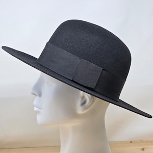 May include: A black fedora hat with a wide brim and a black grosgrain ribbon band. The hat is displayed on a white mannequin head, showcasing its shape and design. The hat's brim extends approximately 3 inches.