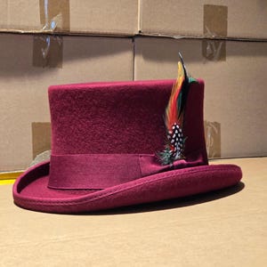 May include: A deep burgundy felt top hat featuring a matching grosgrain ribbon band around the base of the crown. A decorative feather accent with black, white, red, yellow, and green colours is tucked into the side of the ribbon band.