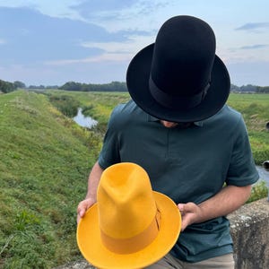 May include: A person wearing a green polo shirt holds a yellow fedora hat in their hands. The hat has a brown ribbon band around the crown. The person is wearing a black fedora hat on their head.