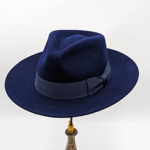 May include: A navy blue fedora hat with a wide brim and a matching grosgrain ribbon band. The hat is displayed on a wooden hat stand against a white background. The hat's crown is indented, and the brim is slightly upturned.
