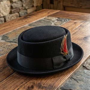 Puede incluir: Un sombrero pork pie negro con copa plana y ala corta levantada. El sombrero está adornado con una cinta de grosgrain negra y un detalle de pluma decorativa en tonos rojos, marrones y beige insertado en el lateral de la cinta.