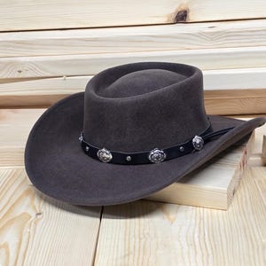 Könnte beinhalten: Ein brauner Cowboyhut aus Filz mit einem schwarzen Lederband und silbernen Conchos. Der Hut hat eine breite Krempe und eine hohe Krone.