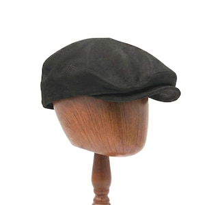 Könnte beinhalten: Eine schwarze Newsboy-Cap aus Wildleder, präsentiert auf einem hölzernen Hutständer. Die Cap hat eine runde Krone und einen kleinen, steifen Schirm. Der Hut ist dunkelgrau und scheint aus weichem Wildleder zu sein.