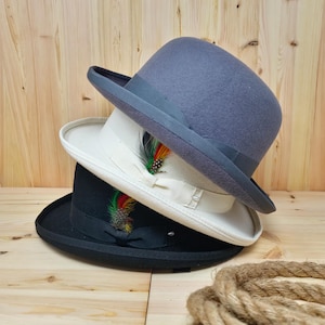 Può includere: Tre cappelli a bombetta impilati uno sopra l'altro. Il cappello superiore è grigio, il cappello intermedio è bianco e il cappello inferiore è nero. Ogni cappello ha una sottile fascia nera attorno alla corona. Il cappello intermedio ha una piccola piuma con accenti rossi, gialli e verdi.