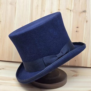 May include: A navy blue top hat with a black ribbon band and bow. The hat is displayed on a brown hat stand. The hat has a classic, formal design.