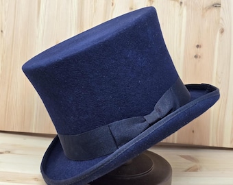 May include: A navy blue top hat with a black ribbon band and bow. The hat is displayed on a brown hat stand. The hat has a classic, formal design.
