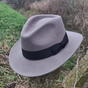 Op de afbeelding: Een grijze fedora hoed met een zwarte band. De hoed is gemaakt van een zacht materiaal en heeft een klassiek ontwerp. De hoed rust buiten op een stenen paal. De rand van de hoed heeft een diameter van ongeveer 30 cm.