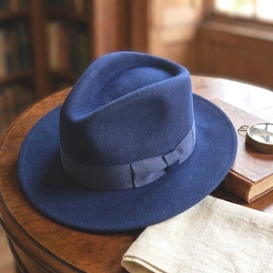 Pu&ograve; includere: Un cappello fedora in feltro blu navy profondo caratterizzato da una corona strutturata con piega centrale e tesa larga. Il cappello &egrave; arricchito da un nastro in gros-grain blu navy abbinato che include un piccolo fiocco piatto sul lato.