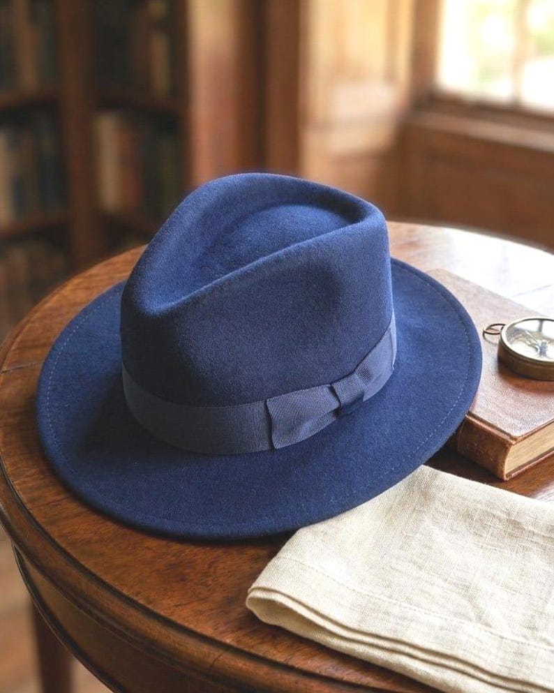 Pu&ograve; includere: Un cappello fedora in feltro blu navy profondo caratterizzato da una corona strutturata con piega centrale e tesa larga. Il cappello &egrave; arricchito da un nastro in gros-grain blu navy abbinato che include un piccolo fiocco piatto sul lato.