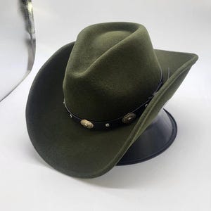May include: An olive green felt cowboy hat featuring a pinched crown and a wide, curved brim. A black leather band encircles the base of the crown, decorated with small, circular, metallic silver-toned studs and ornate, embossed floral-patterned conchos.