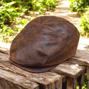 Puede incluir: Una gorra plana de cuero marrón con un acabado desgastado. La gorra tiene una corona redonda y una visera corta.